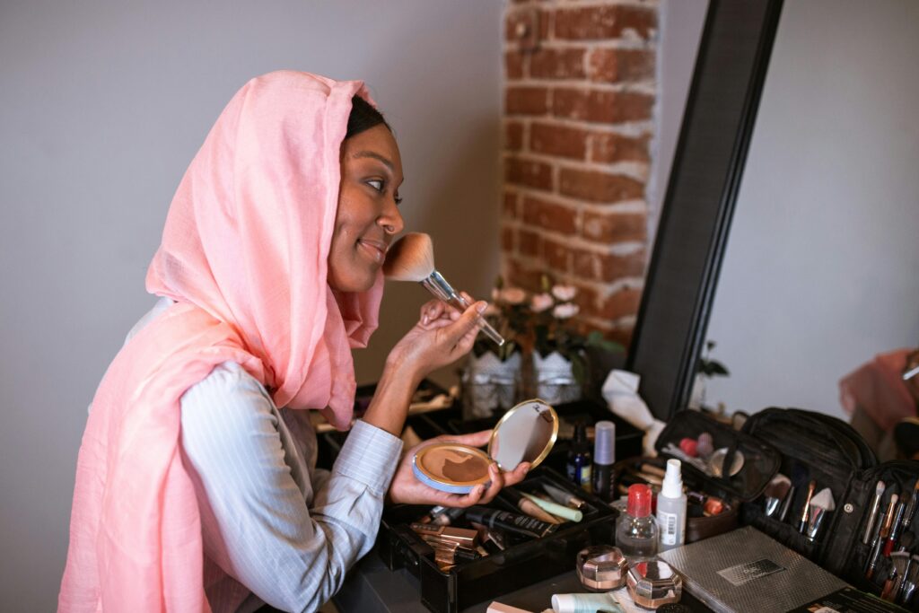 A woman in a pink hijab applying makeup in front of a vanity mirror indoors.