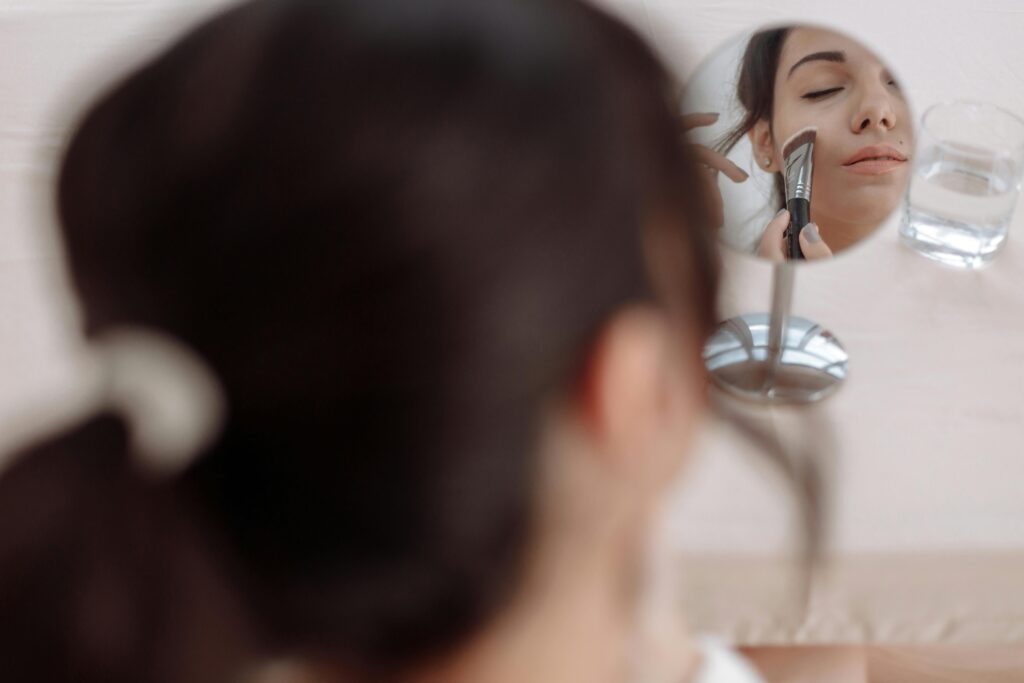 A woman applying makeup using a brush while looking into a small mirror.